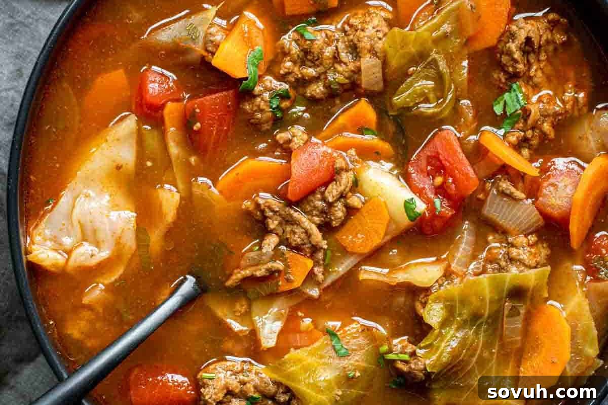 A close-up view of hearty vegetable soup with chunks of tomatoes, carrots, cabbage, ground meat, and a spoon in the bowl.