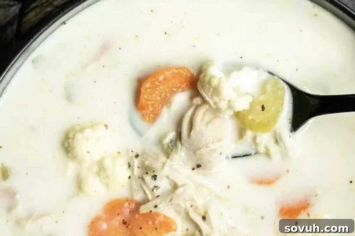 A bowl of creamy white soup with vegetables like carrots, cauliflower, and celery, and pieces of chicken. A spoon is lifting some of the soup.