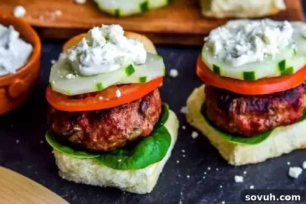 Two slider sandwiches with grilled meat patties, spinach leaves, tomato slices, cucumber slices, and a dollop of white sauce, on a square bread base on a dark surface.