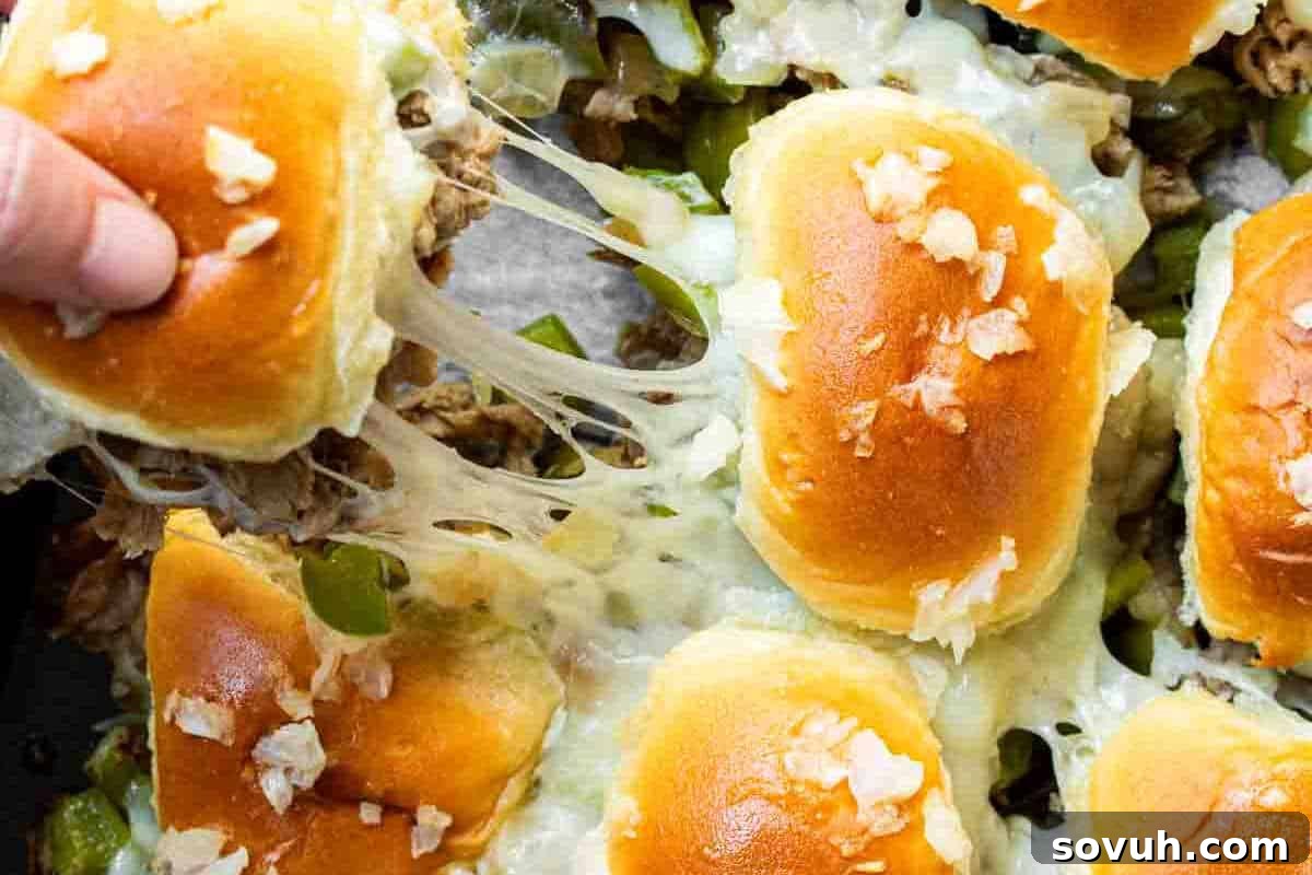A person pulls a slider topped with melted cheese and onions from a tray of prepared sliders. The buns appear golden brown and the filling includes green peppers and cheese.