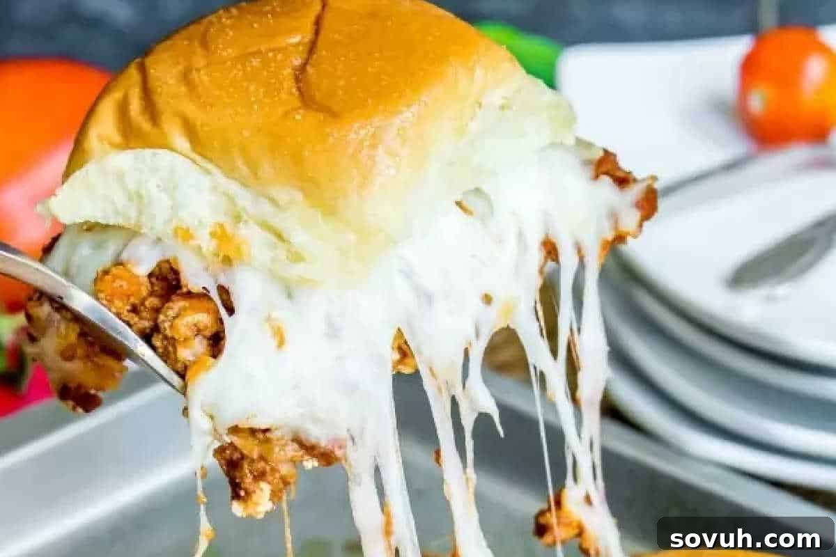 A close-up of a cheesy slider being lifted from a tray, with gooey melted cheese stretching from the bun. Plates and cutlery are visible in the background.