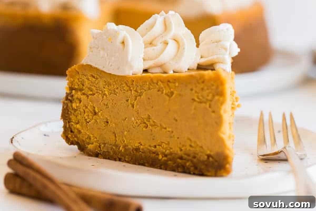 A generous slice of creamy pumpkin cheesecake, artfully topped with a dollop of fluffy whipped cream, sits invitingly on a white plate beside a gleaming silver fork. Two aromatic cinnamon sticks are elegantly placed beside the plate, adding a festive autumn touch.