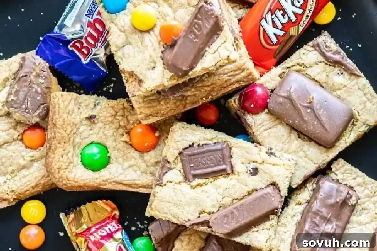 Decadent cookie bars generously topped with a medley of chopped chocolate pieces and colorful Halloween candies, beautifully surrounded by various wrapped chocolate bars, making a spectacular dessert.