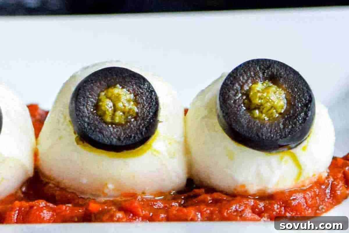 Smooth mozzarella balls artfully topped with slices of black olives and intricate red pesto 'veins', resting on a bed of vibrant tomato sauce, creating deliciously creepy Halloween eyeballs.