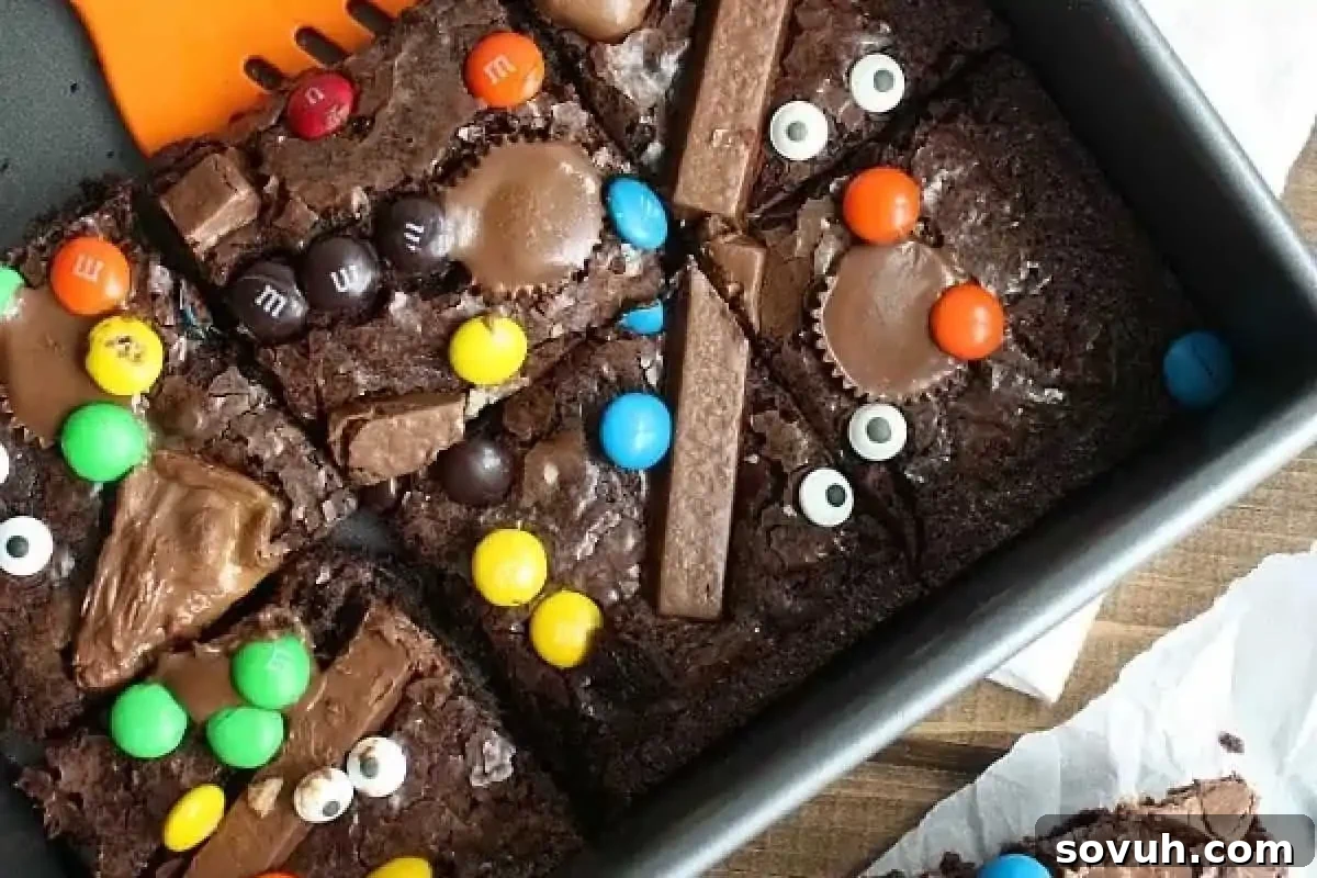A rich baking pan filled with decadent chocolate brownies, generously topped with a colorful assortment of Halloween candies like chocolate pieces, M&M's, and peanut butter cups, creating a 'Witch's Brew' effect.