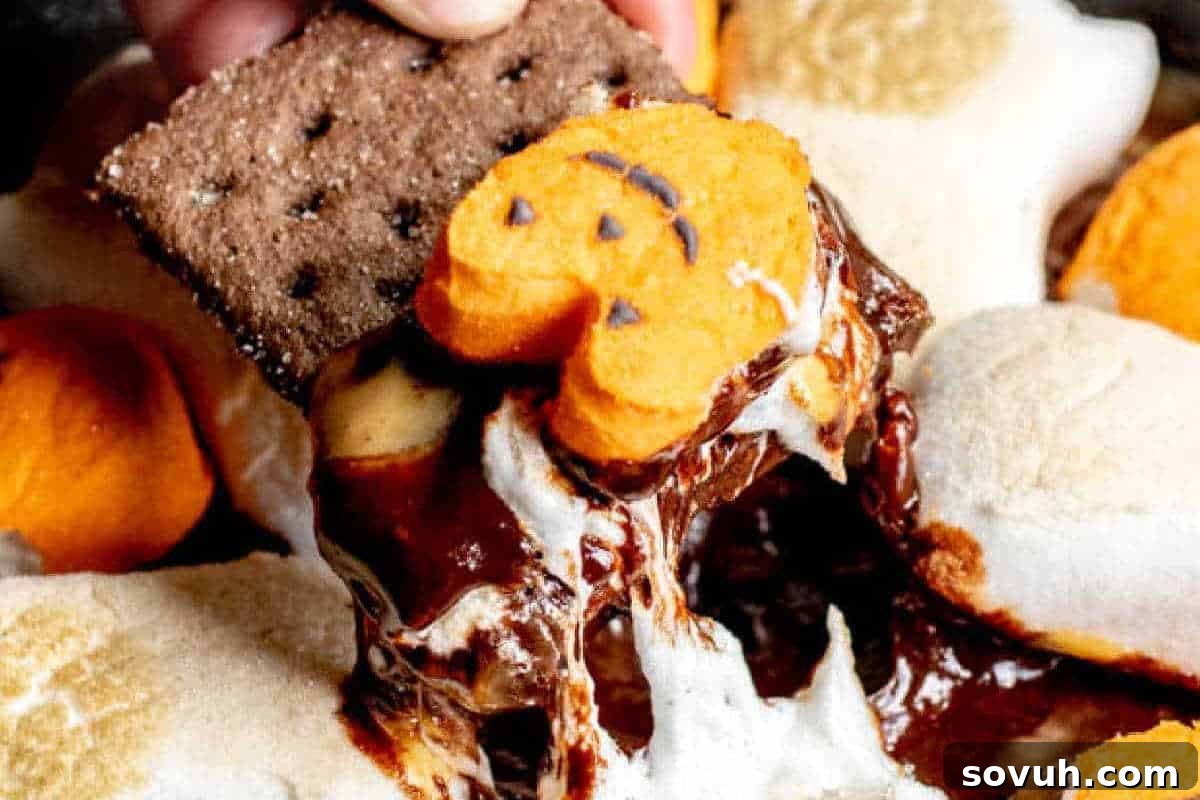 A hand dipping a graham cracker into a gooey, melted Halloween S'mores Dip, topped with a charming pumpkin-shaped marshmallow, creating a perfect warm and sweet seasonal treat.