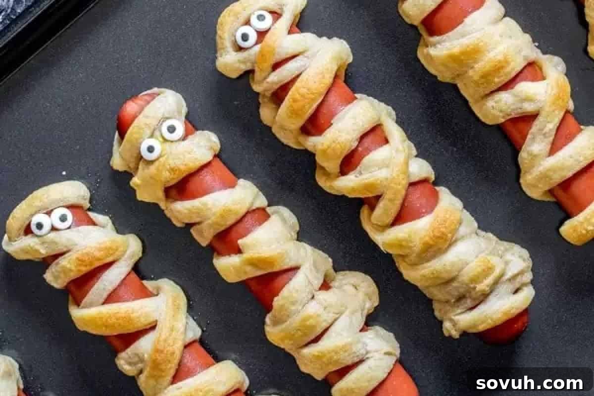 Four delicious hot dogs wrapped in golden pastry strips to resemble playful mummies, with two featuring edible candy eyes, resting on a dark, rustic surface, ideal for a Halloween party appetizer.