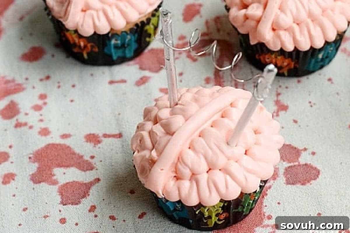 Moist cupcakes topped with realistic, pink brain-shaped icing, complete with ridges and grooves, and finished with clear, glistening decorations, all arranged on a splattered tablecloth for a creepy effect.