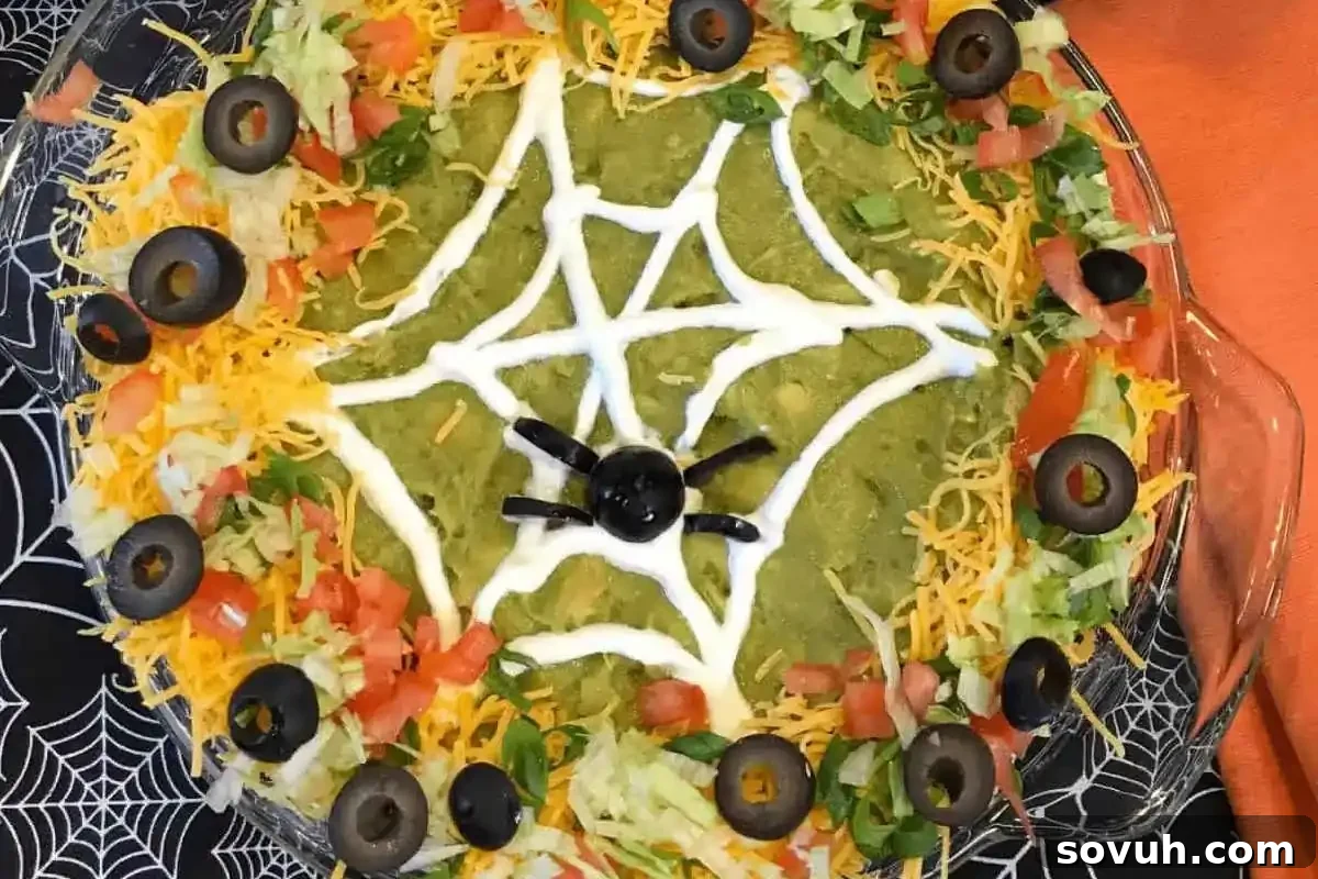 A layered dip with guacamole, shredded cheese, lettuce, tomatoes, and olives, decorated with a sour cream spider web and an olive spider on top.
