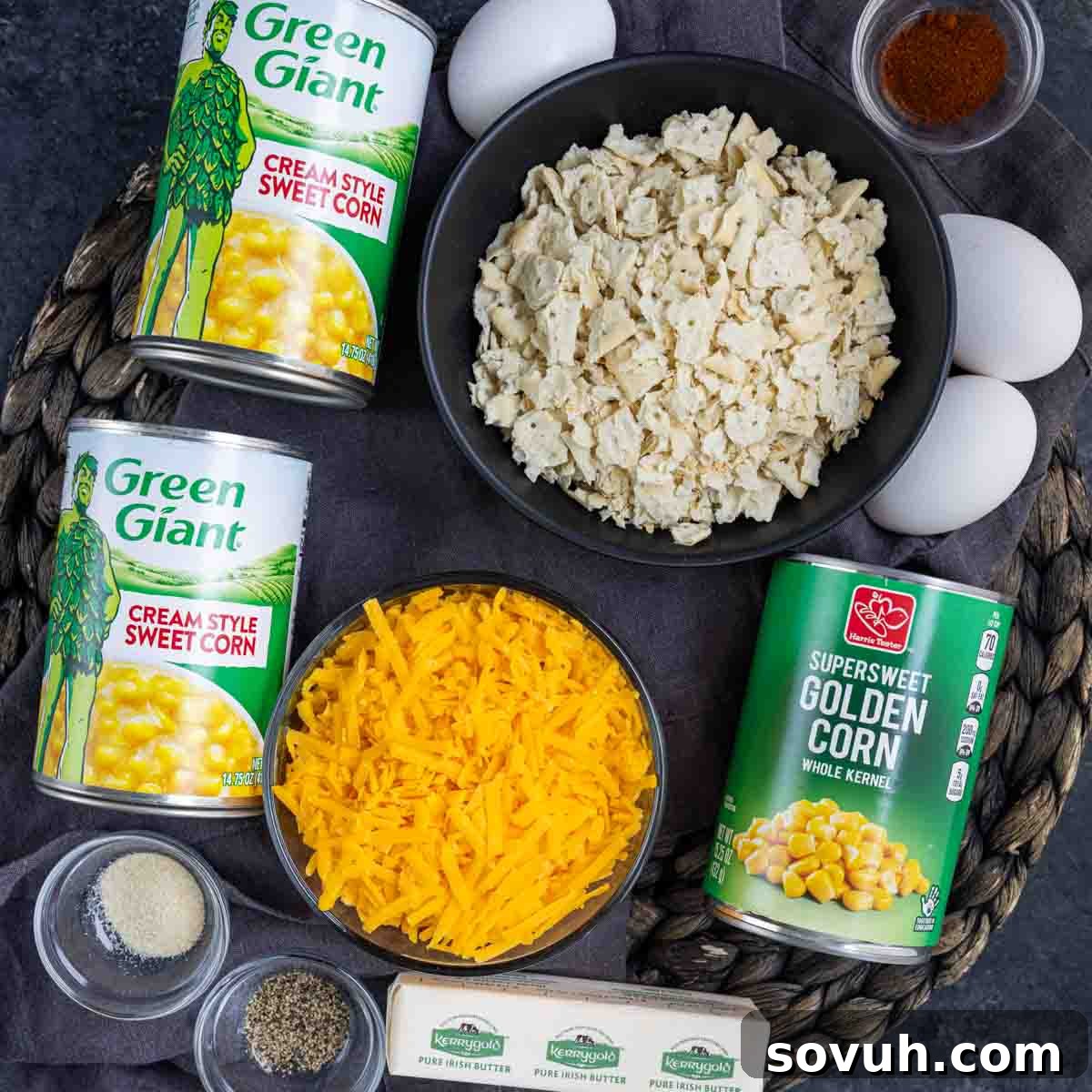 Ingredients for a corn casserole recipe, including cans of cream style corn, whole kernel corn, eggs, crushed crackers, shredded cheese, butter, and seasonings, arranged on a dark surface.