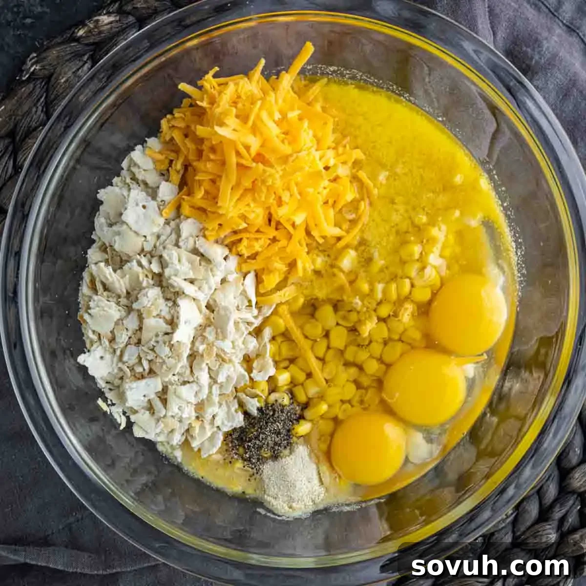 A glass bowl containing shredded cheese, corn, eggs, crushed crackers, and seasoning.