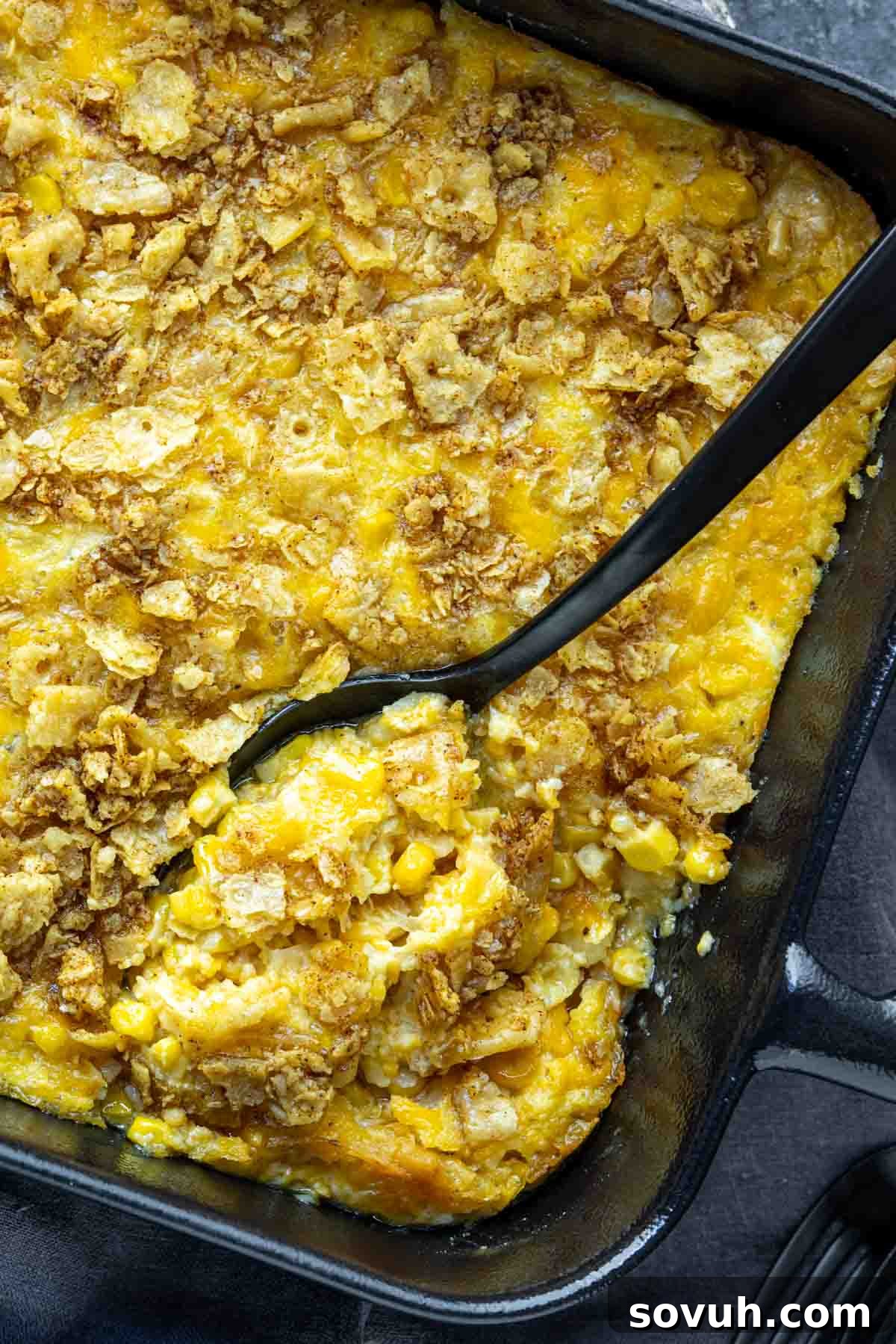 A baking dish filled with cheesy corn casserole topped with crushed crackers, with a serving spoon partially removing a portion.