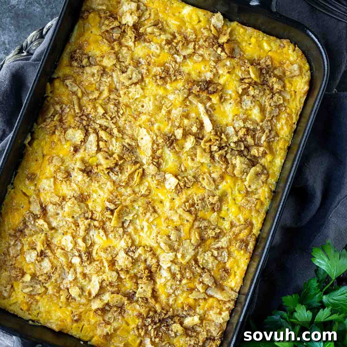 A baked casserole with a golden, crispy topping in a rectangular dish, placed on a dark surface beside fresh parsley.