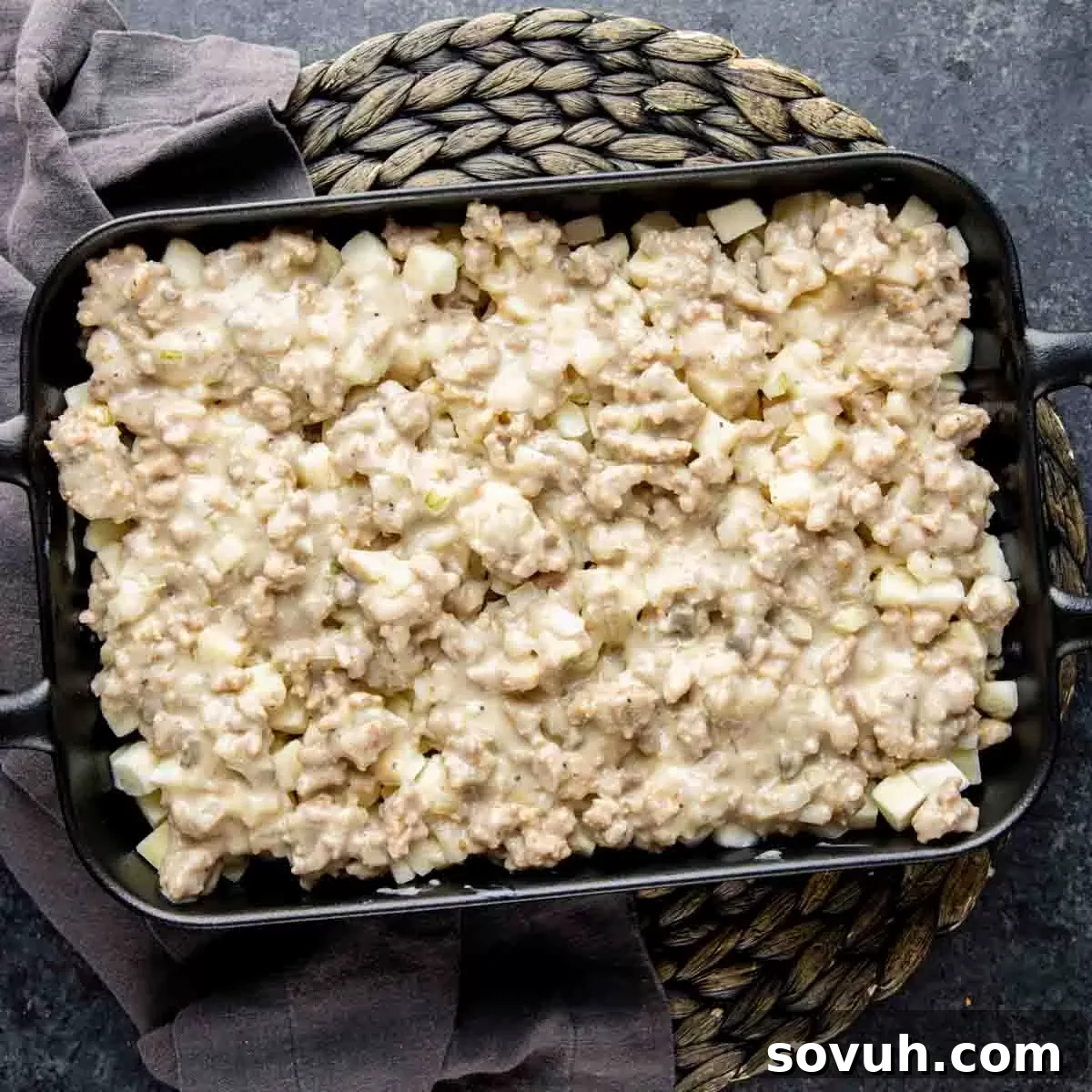 A baking dish half-filled with layers of frozen diced potatoes, creamy sausage mixture, and shredded cheddar cheese, ready for the oven.