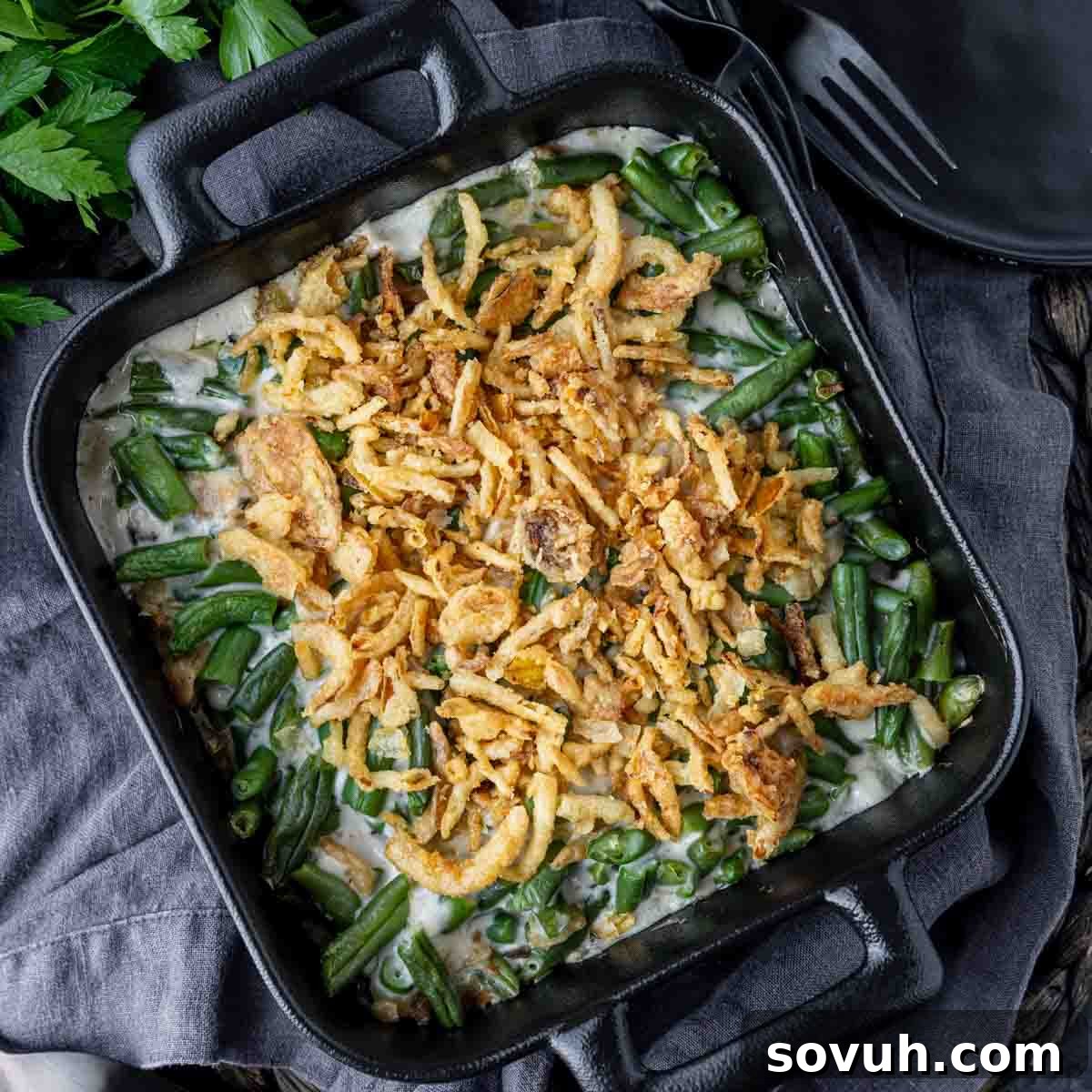 Square dish with green bean casserole topped with crispy fried onions, placed on a dark cloth.
