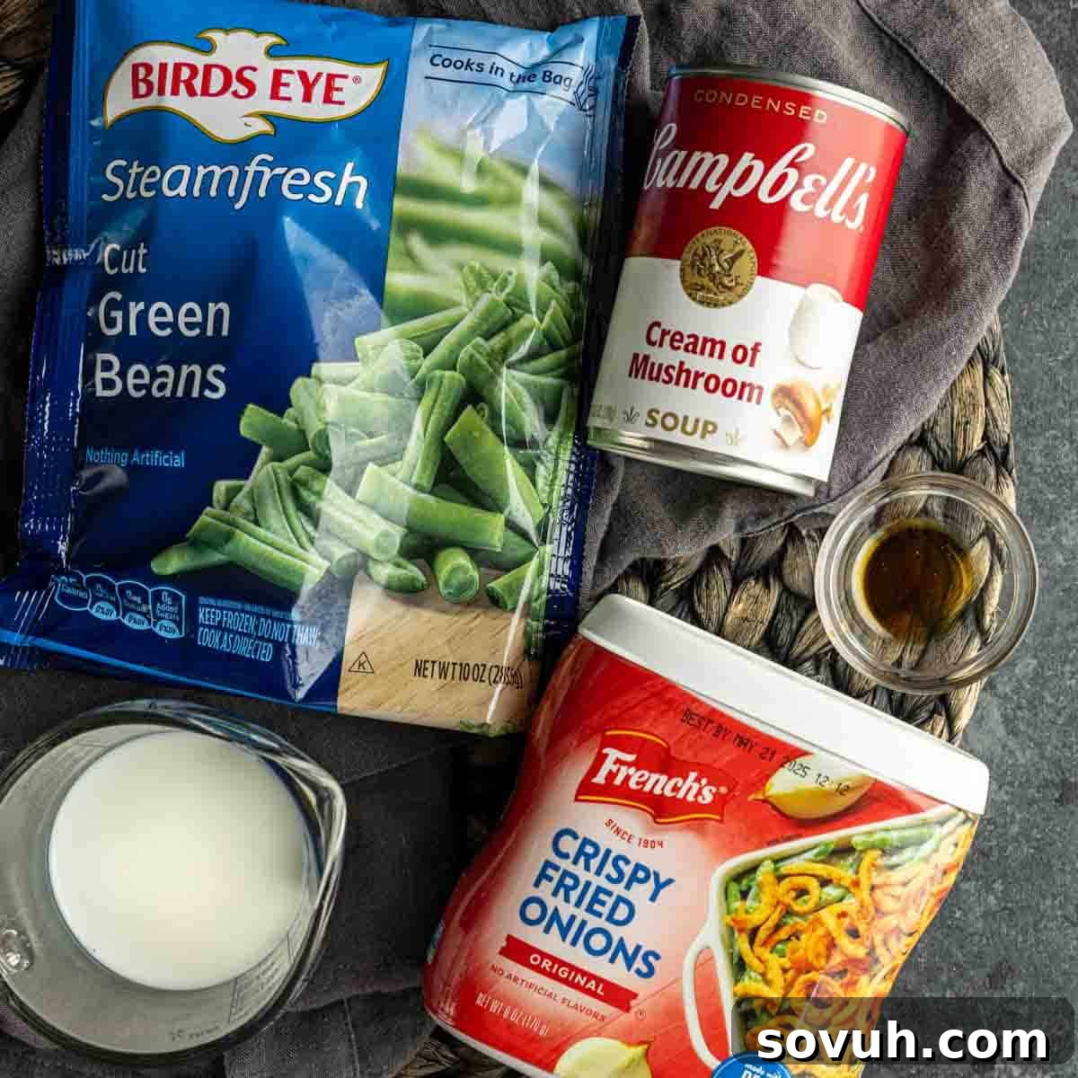 Ingredients for a green bean casserole including a bag of frozen green beans, can of cream of mushroom soup, container of crispy fried onions, a glass of milk, and a small dish of liquid.