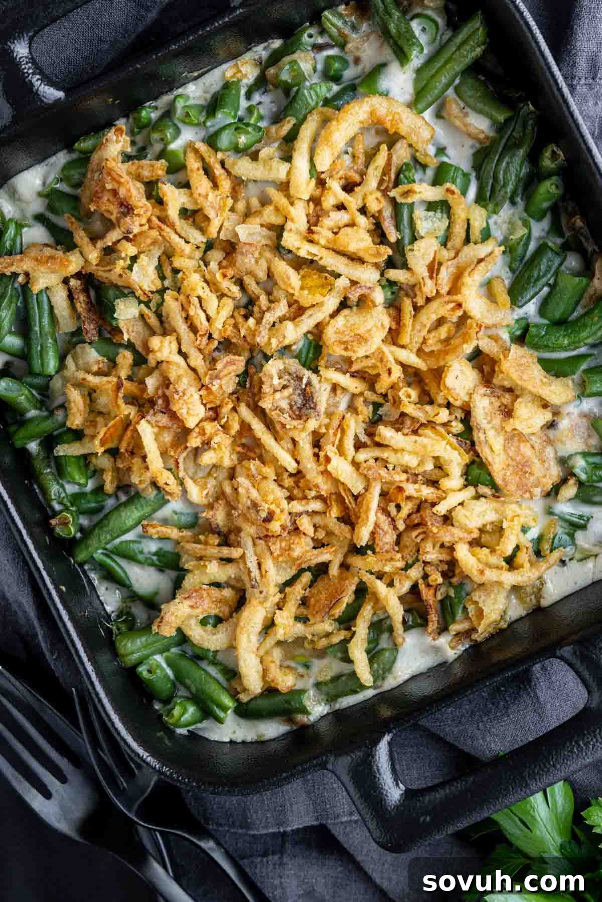 A baked green bean casserole topped with crispy fried onions in a black baking dish, placed on a dark cloth with parsley and fork nearby.
