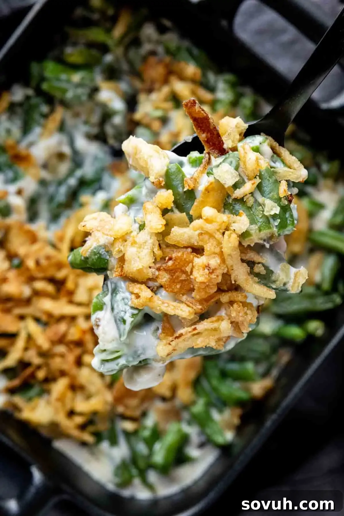 A spoonful of green bean casserole topped with crispy fried onions is lifted from a baking dish.