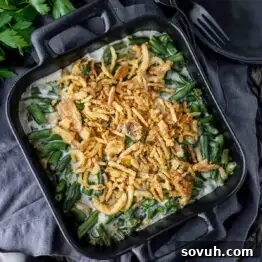Square dish with green bean casserole topped with crispy fried onions, placed on a dark cloth.