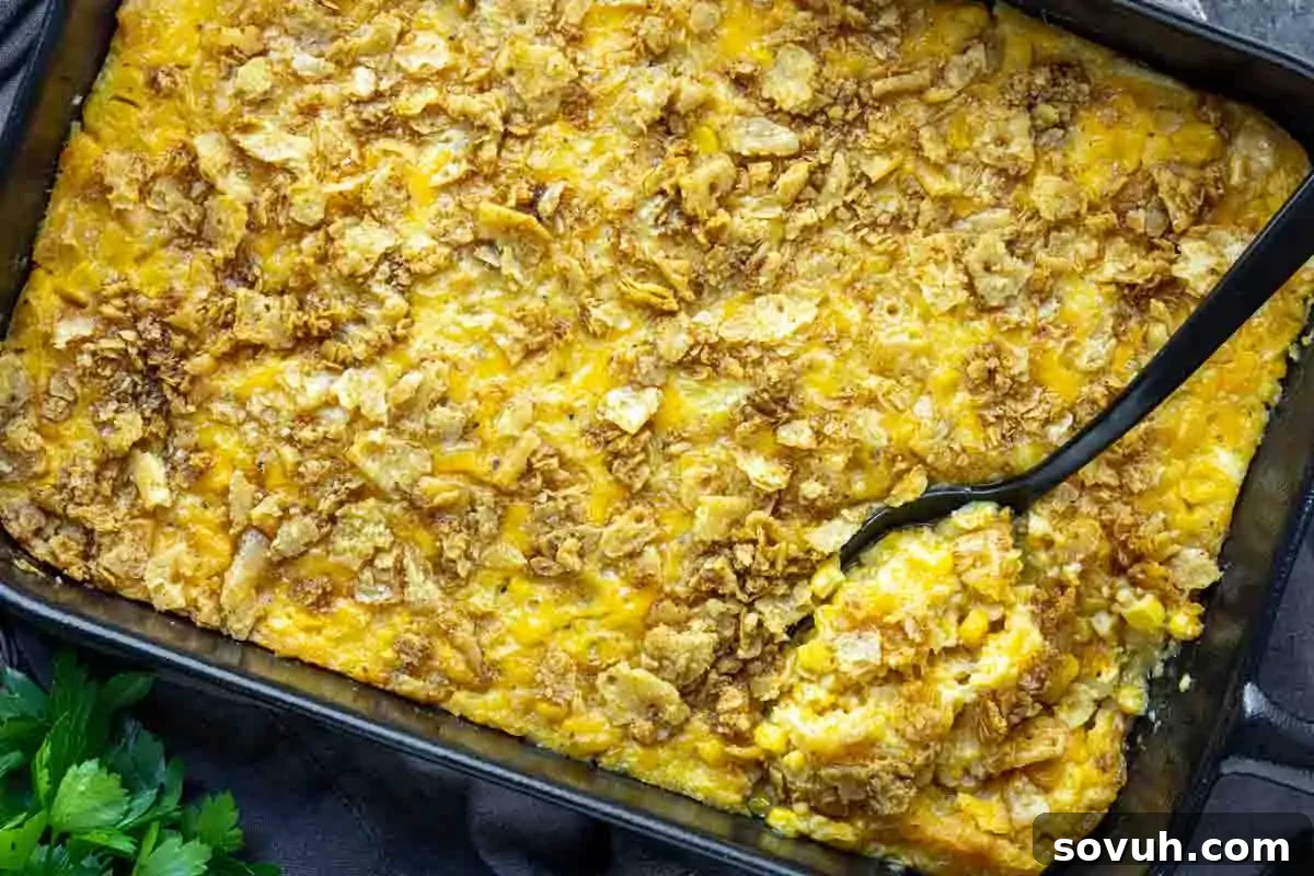 A rectangular baking dish filled with golden-brown Scalloped Corn casserole, topped with a crisp layer of crushed crackers and melted cheese, with a black serving spoon.