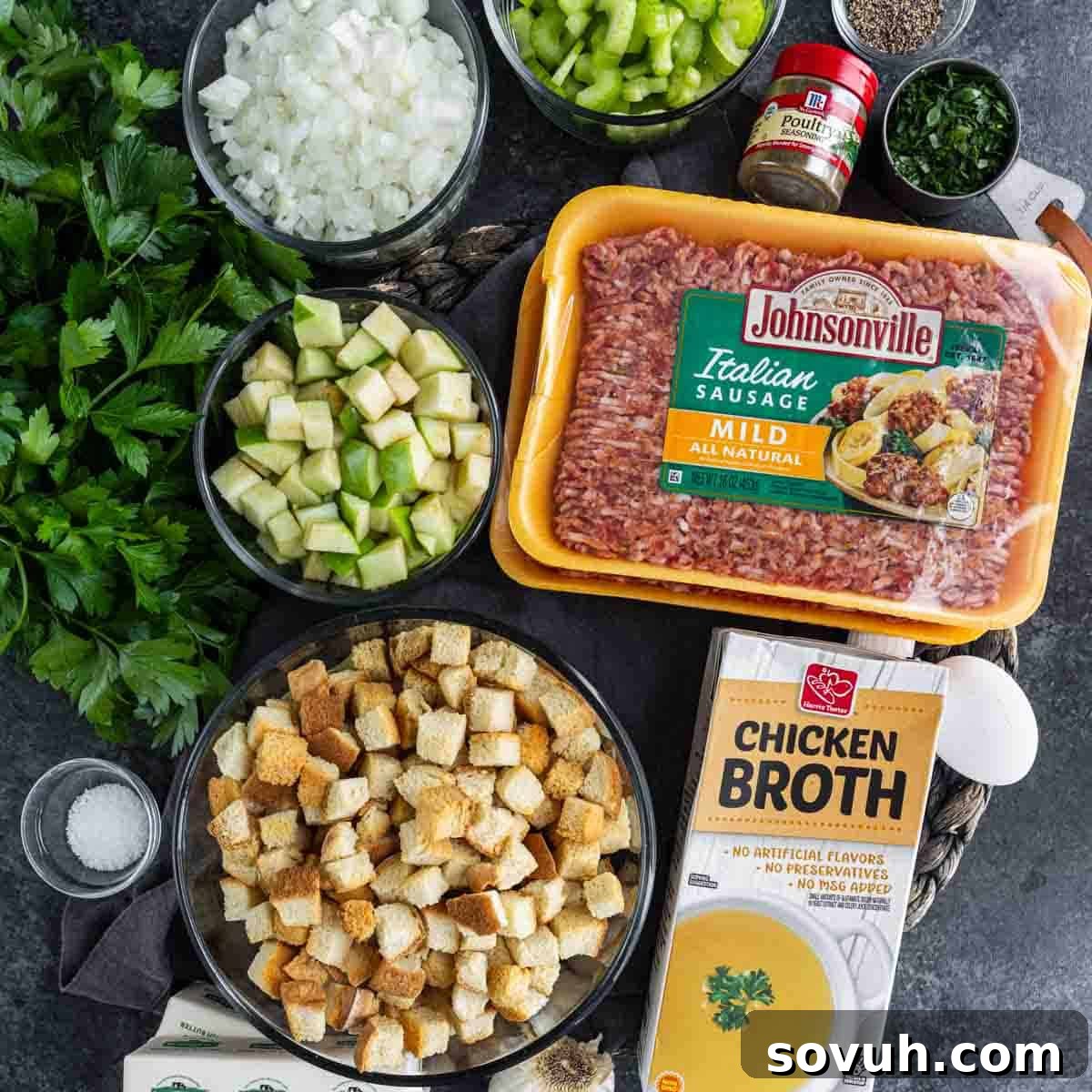 Ingredients for a stuffing recipe: Italian sausage, chicken broth, cubed bread, chopped onions, celery, parsley, zucchini, spices, and salt on a dark surface.