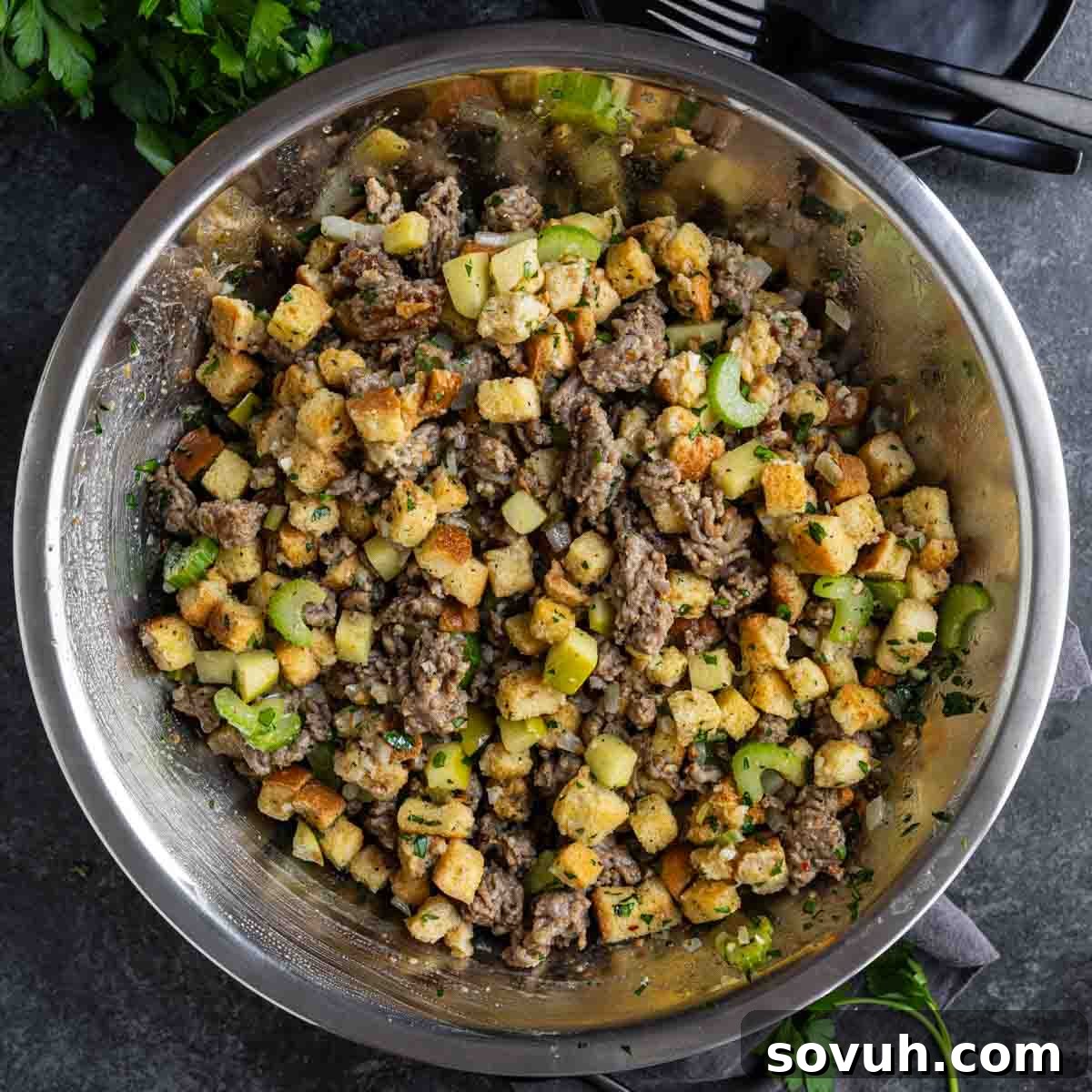 A large stainless steel bowl filled with a stuffing mixture of diced bread, cooked ground meat, chopped celery, and herbs.