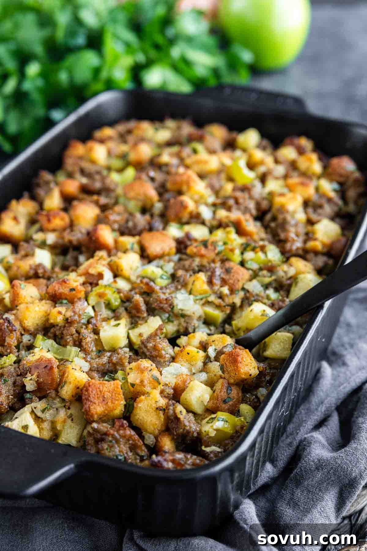 A baking dish filled with golden brown stuffing, featuring cubes of bread, pieces of sausage, and green herbs. A black serving spoon is visible in the dish.