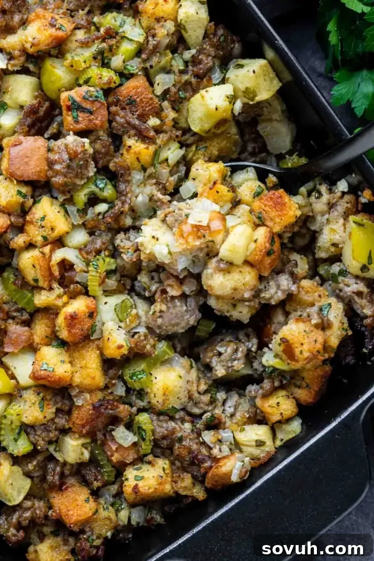 A tray of stuffing with cubes of bread, diced celery, onions, sausage, and herbs, with a serving spoon resting on top.
