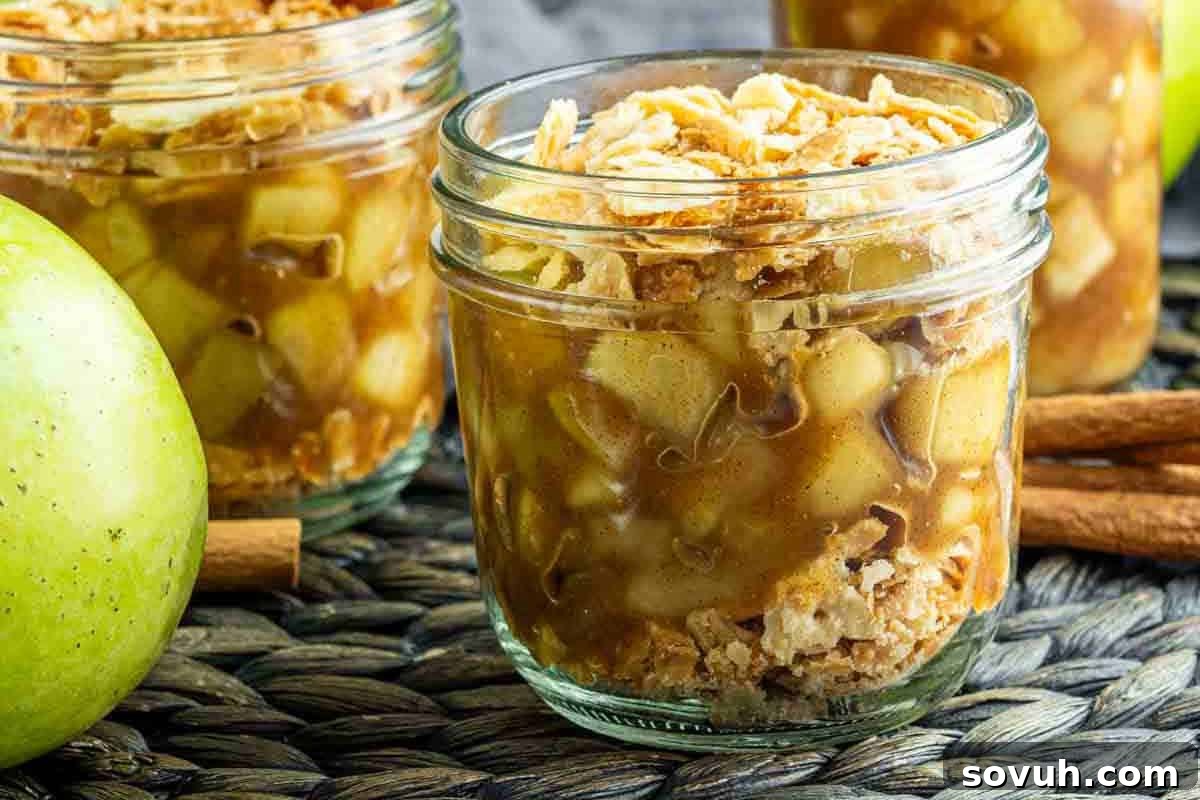 Glass jars filled with apple crisp on a woven surface, with green apples and cinnamon sticks nearby.