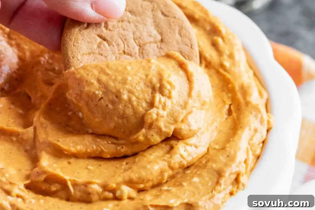 A hand holds a cookie, dipping it into a creamy orange pumpkin dip in a white bowl.