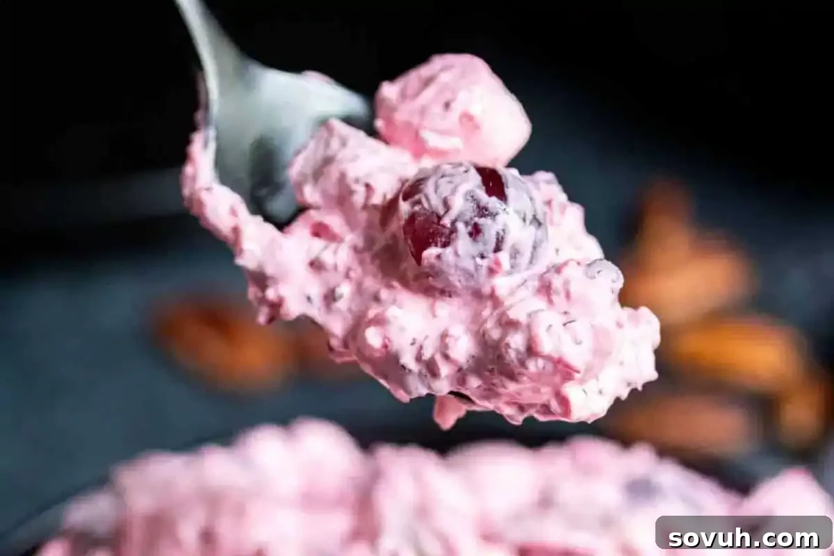 A vibrant spoonful of fluffy pink fruit salad, highlighting a juicy red grape, set against a softly blurred background, suggesting a delightful and refreshing side dish for holiday celebrations.