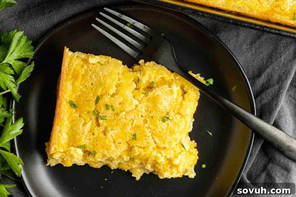 A hearty slice of golden, cheesy cornbread casserole on a sleek black plate, accompanied by a fork and garnished with fresh green herbs, with delicate parsley leaves scattered nearby, showcasing a perfect holiday side.