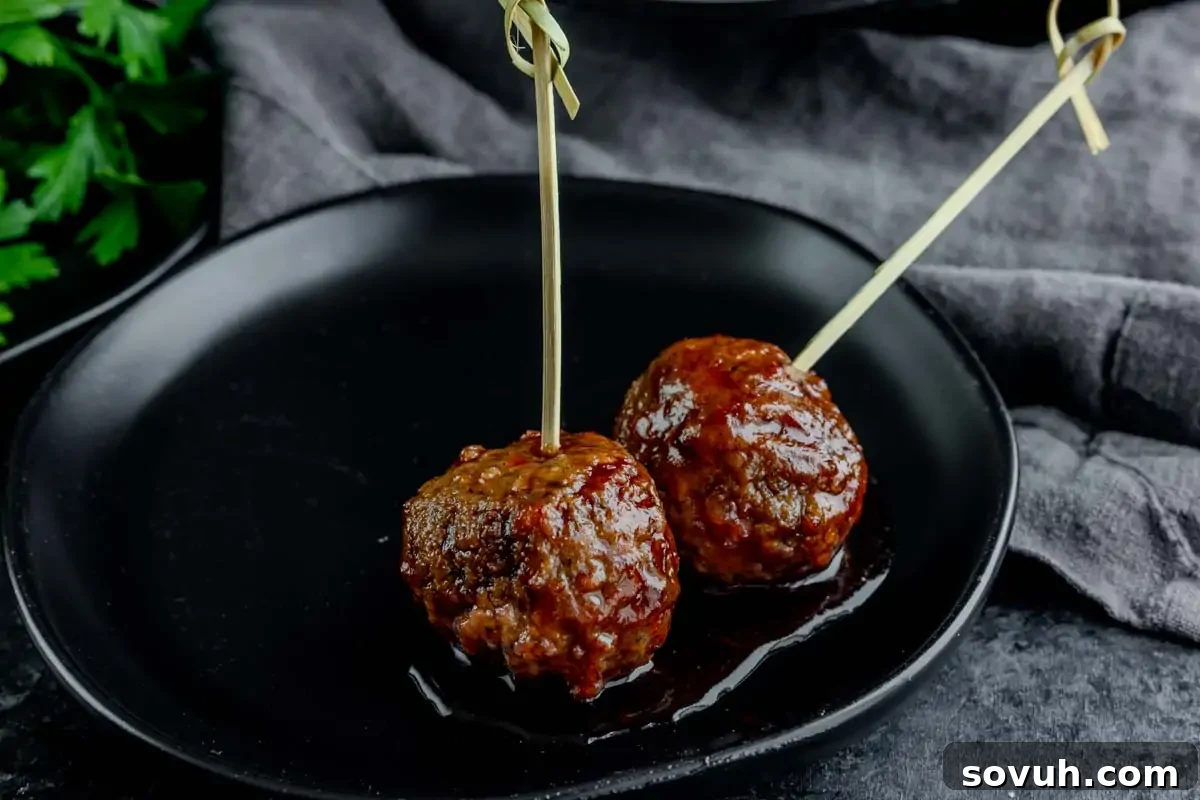 Two glazed meatballs on skewers sit on a black plate, with a dark cloth and green leaves in the background.