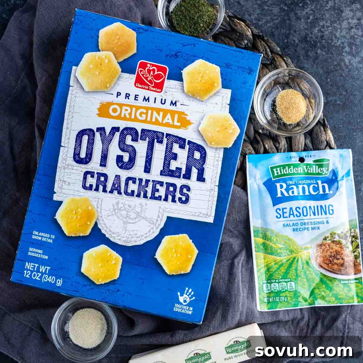 Box of original oyster crackers surrounded by bowls of spices, Hidden Valley Ranch seasoning packet, and a stick of butter on a dark surface.