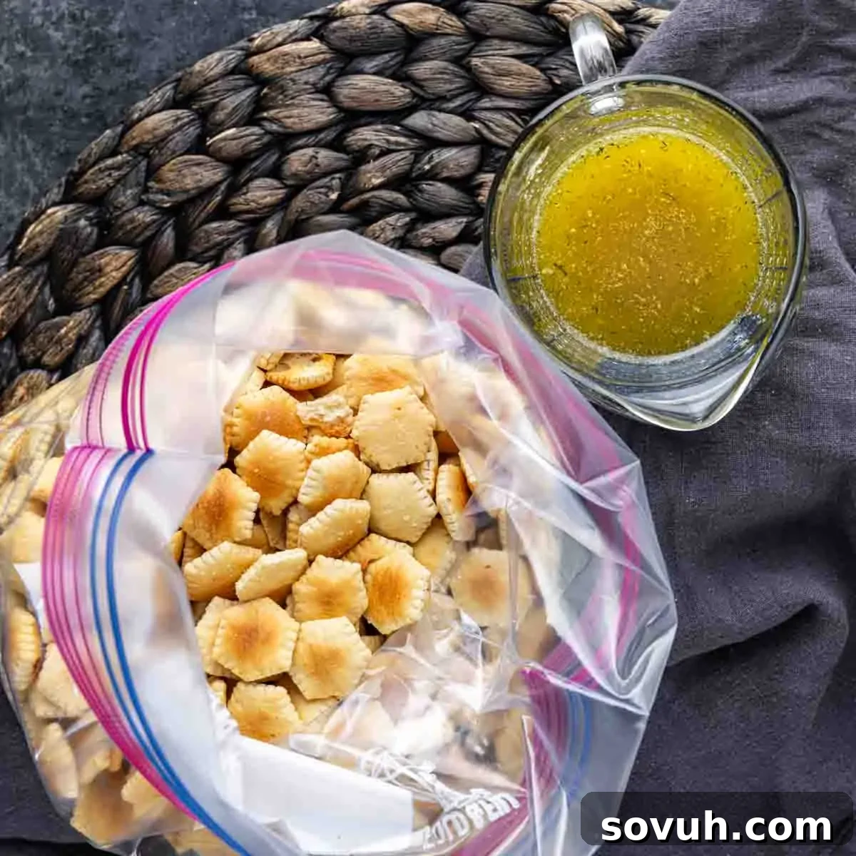 A plastic bag filled with crackers next to a measuring cup containing seasoned oil, placed on a woven mat.