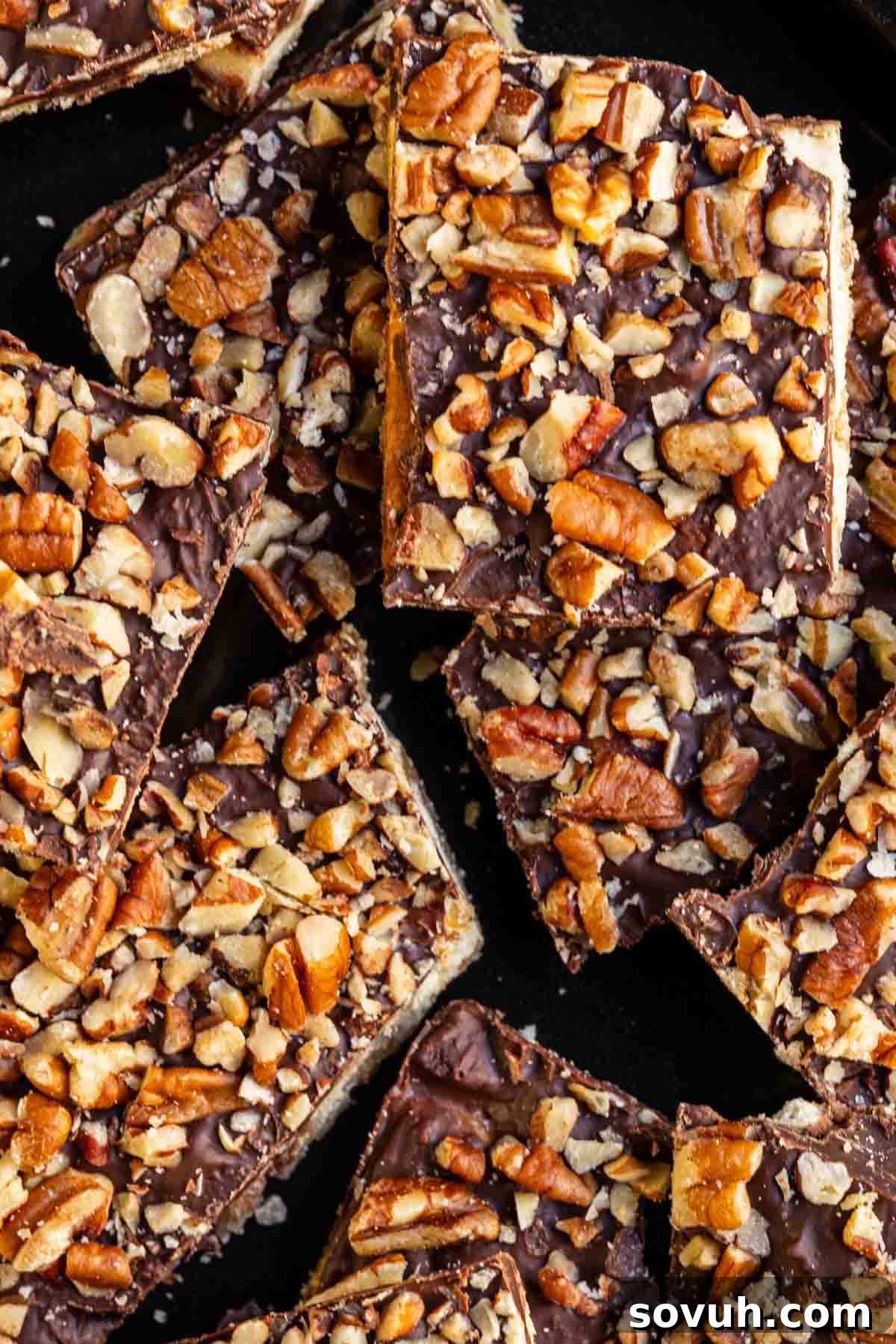 Squares of chocolate toffee bark topped with chopped pecans on a dark surface.