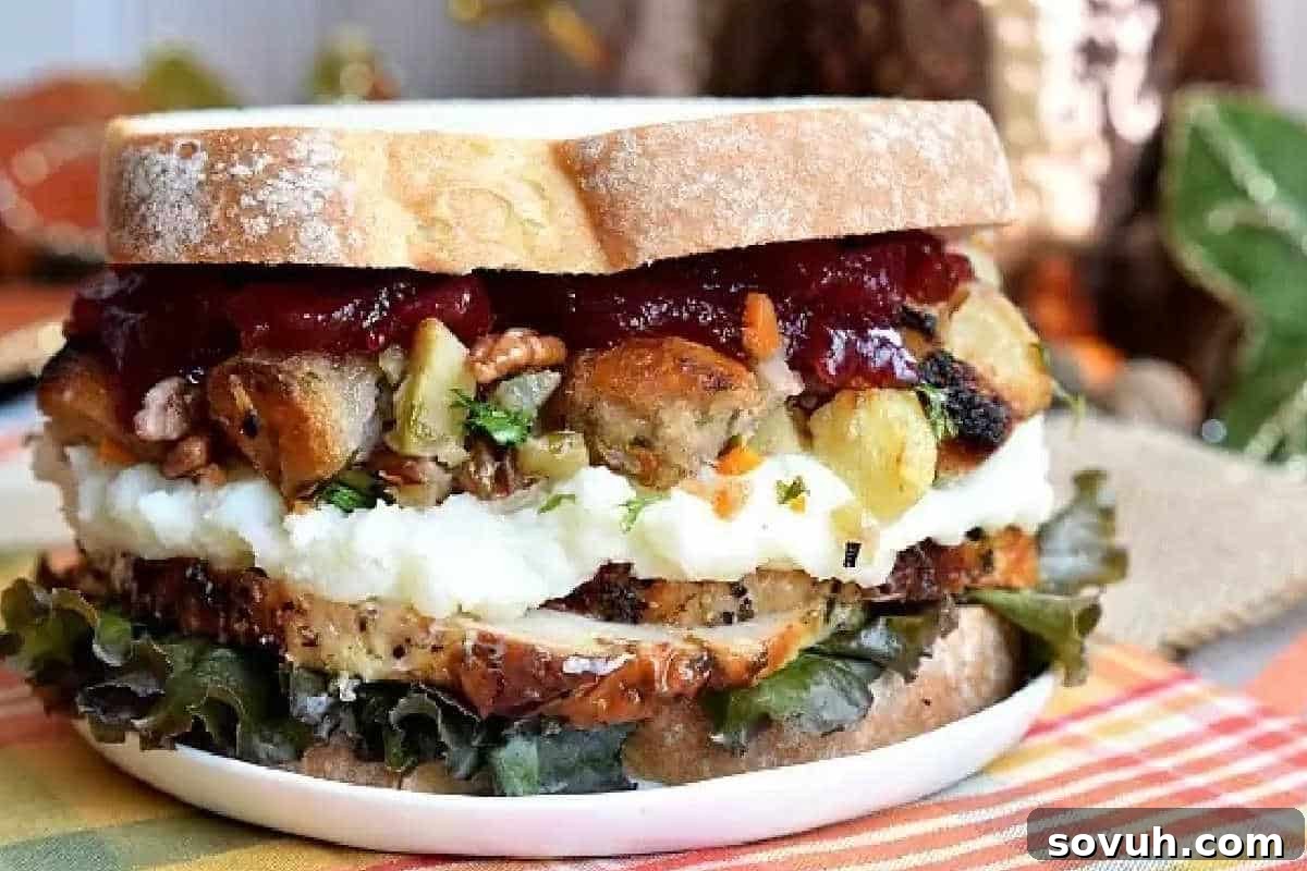 A perfectly constructed Thanksgiving Leftovers Sandwich, featuring thick slices of turkey, savory stuffing, creamy mashed potatoes, tangy cranberry sauce, and crisp lettuce, all nestled between two slices of golden-brown bread on a plate.