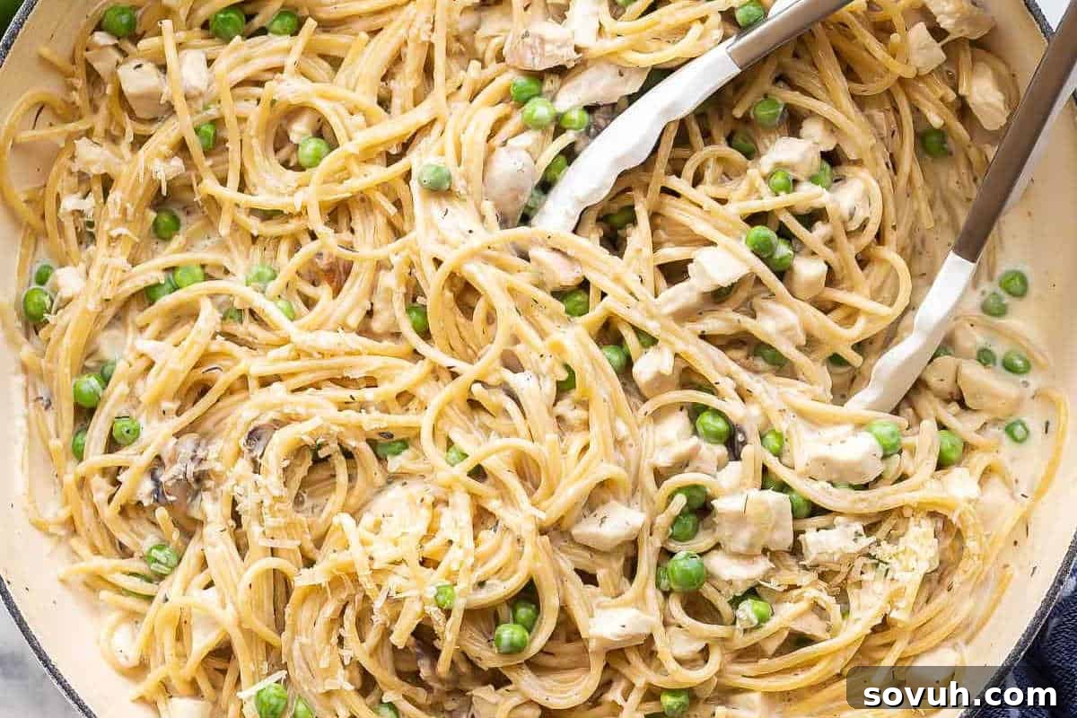 A close-up shot of creamy spaghetti with generous pieces of turkey, tender peas, and sautéed mushrooms, being expertly mixed with tongs in a single pan.