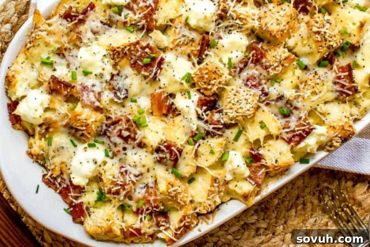 A baked dish with crispy bread pieces, melted cheese, bacon, and chopped chives on top, served on a white oval plate, an Everything Bagel Make Ahead Breakfast Casserole.