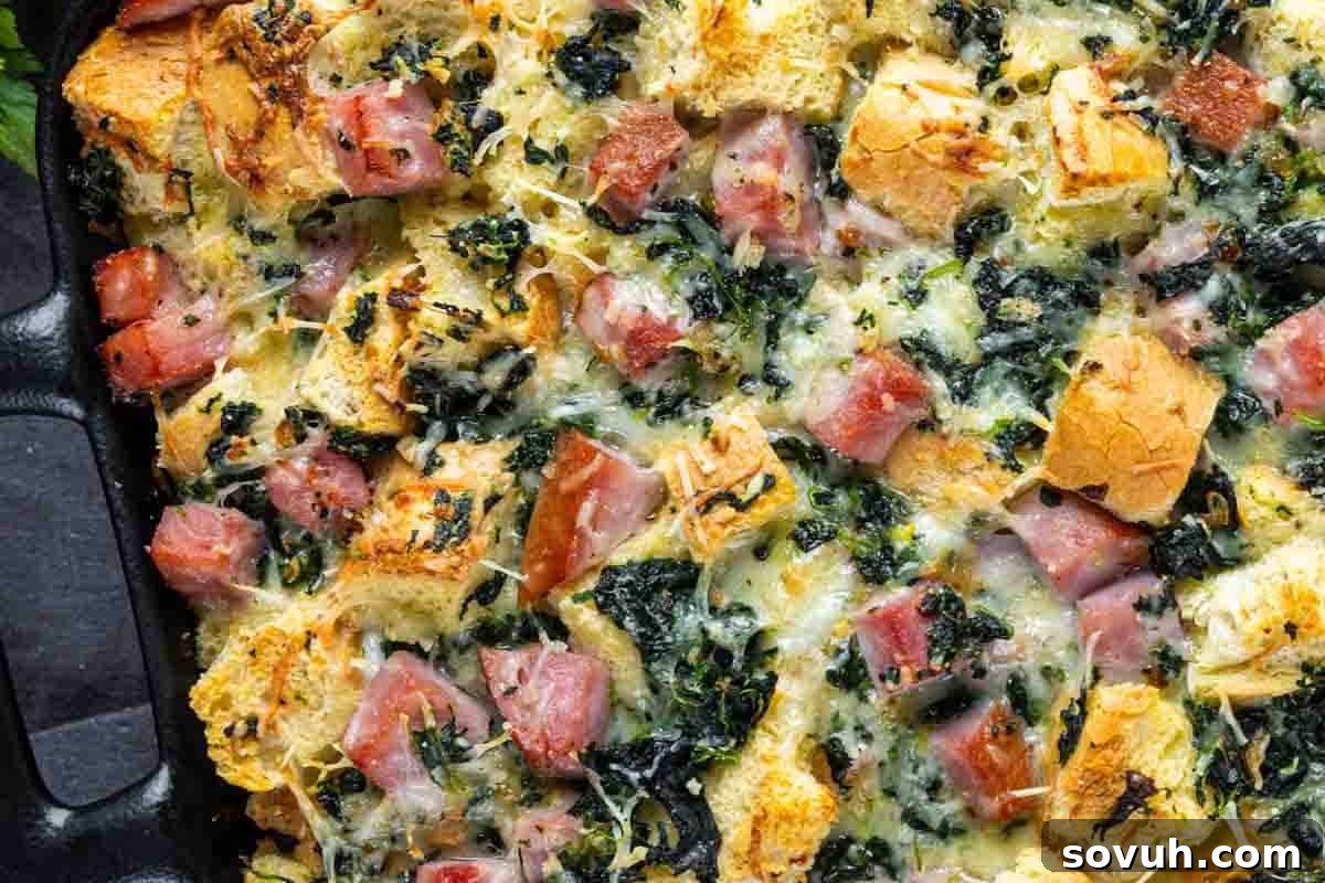 Close-up of a baked casserole with cubed bread, ham, spinach, and melted Gruyère cheese in a black dish, a savory breakfast strata.