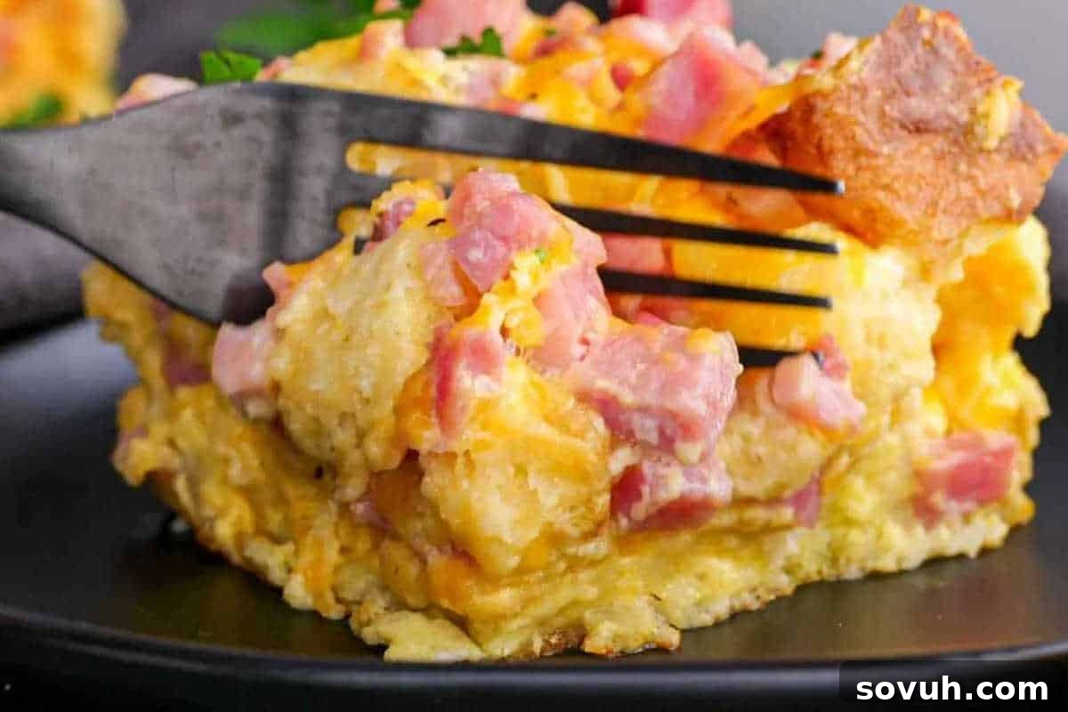 A fork cutting into a slice of ham and cheese breakfast casserole on a black plate, featuring English muffins.