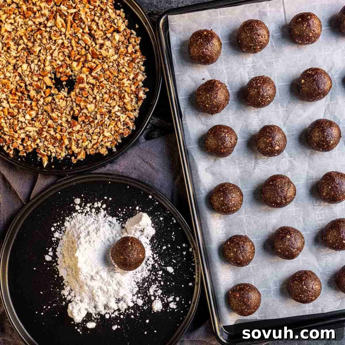 Tray of chocolate balls on parchment paper, surrounded by plates of crushed nuts and powdered sugar. One chocolate ball is partially coated with powdered sugar.
