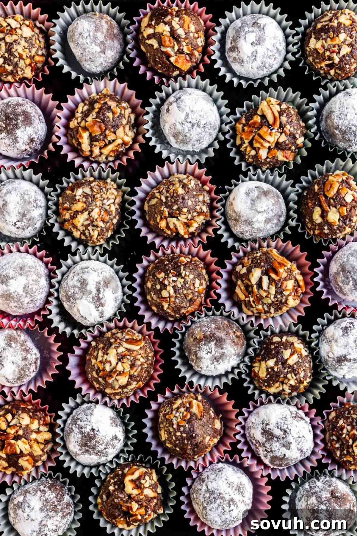 An overhead view of assorted chocolate truffles in paper cups, topped with powdered sugar and chopped nuts, arranged in a grid pattern.