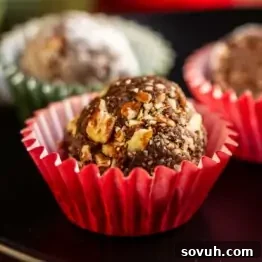 A chocolate truffle coated with chopped nuts sits in a red paper cup, with other truffles blurred in the background.