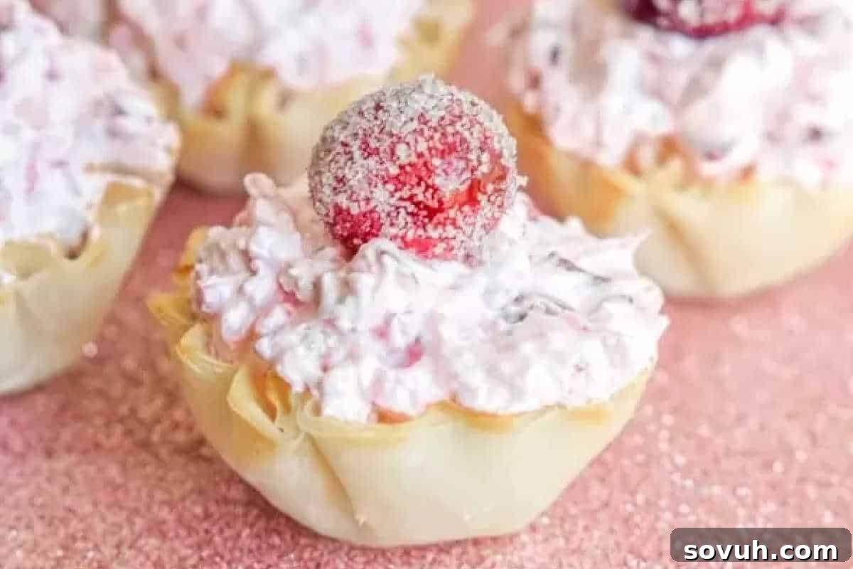 Delicate mini pastry cups, brimming with a light and airy pink cranberry fluff mixture, each elegantly crowned with a glistening, sugar-coated cherry, all arranged on a festive, subtly glittery pink surface.