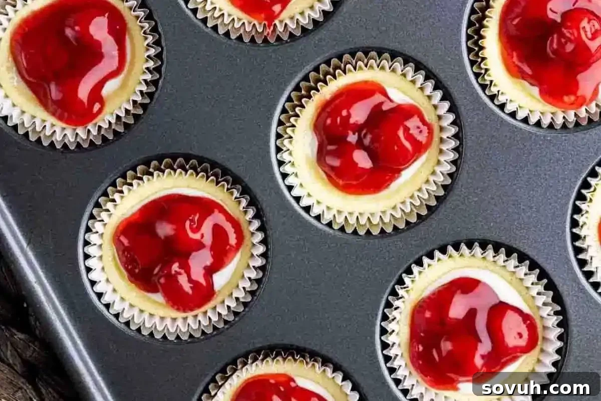 Delightful mini cheesecakes, perfectly portioned in a muffin tray, each sumptuously topped with a glistening ruby-red cherry filling, presenting a charming and irresistible dessert option.