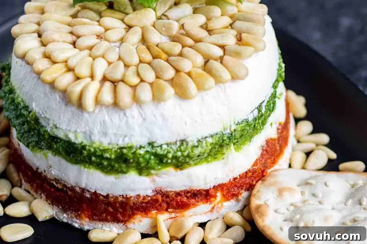 A beautifully layered cheese terrine featuring vibrant green pesto and rich sun-dried tomato layers, elegantly topped and surrounded by crunchy pine nuts, positioned invitingly next to a crisp cracker on a sophisticated black plate.