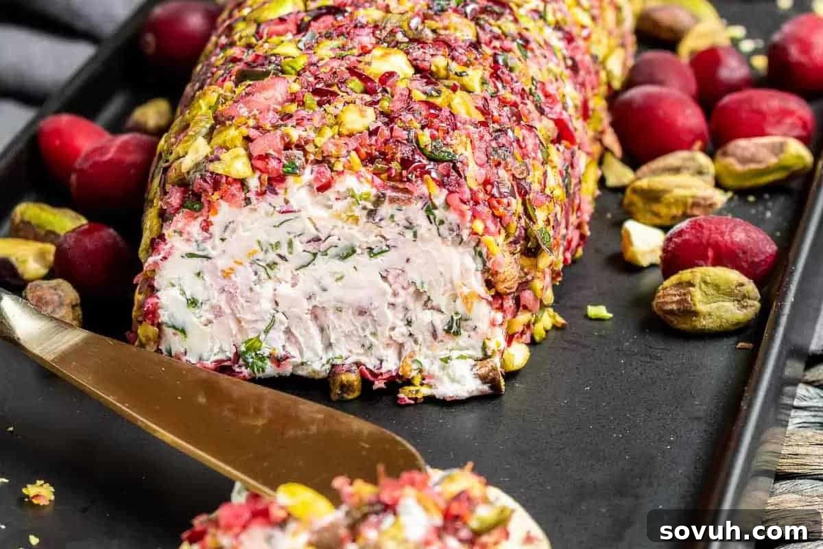 A vibrant goat cheese log, beautifully coated with a mosaic of finely chopped pistachios and jewel-toned cranberries, artfully arranged on a serving tray with scattered fresh cranberries and whole pistachios, next to a golden cheese spreader for elegant serving.