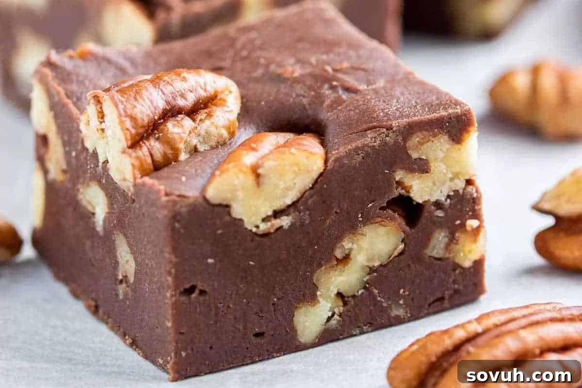 A mouth-watering close-up of thick, rich chocolate fudge squares, featuring whole pecan nuts embedded into their smooth, glossy surface, resting on a pristine white background.