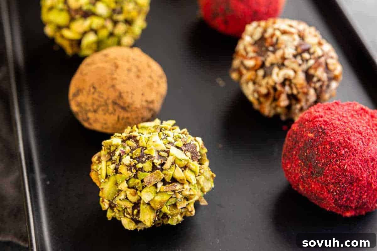 A stunning arrangement of assorted dark chocolate truffles, each exquisitely coated in a variety of toppings including finely chopped pistachios, rich cocoa powder, crunchy crushed nuts, and a dusting of vibrant red powder, artfully presented on a sleek black surface.